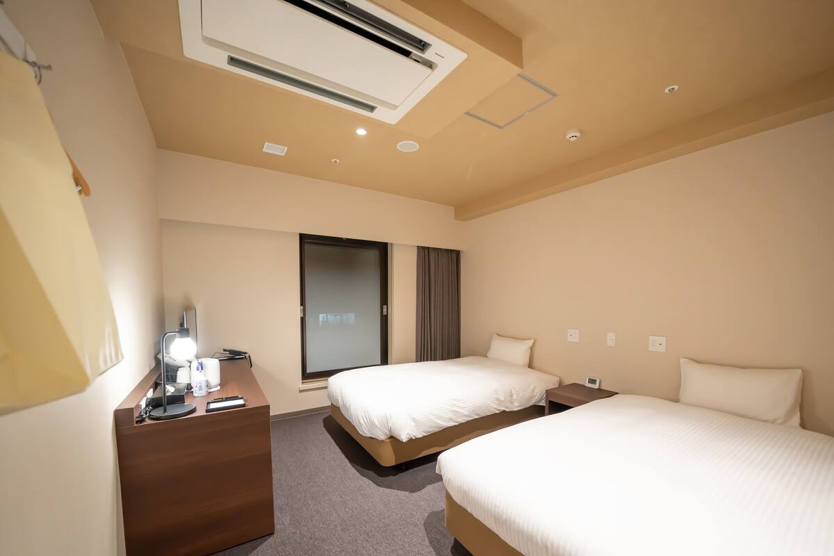 A hotel room with two beds, a desk with a lamp, a window with curtains, and an air conditioning unit on the ceiling.
