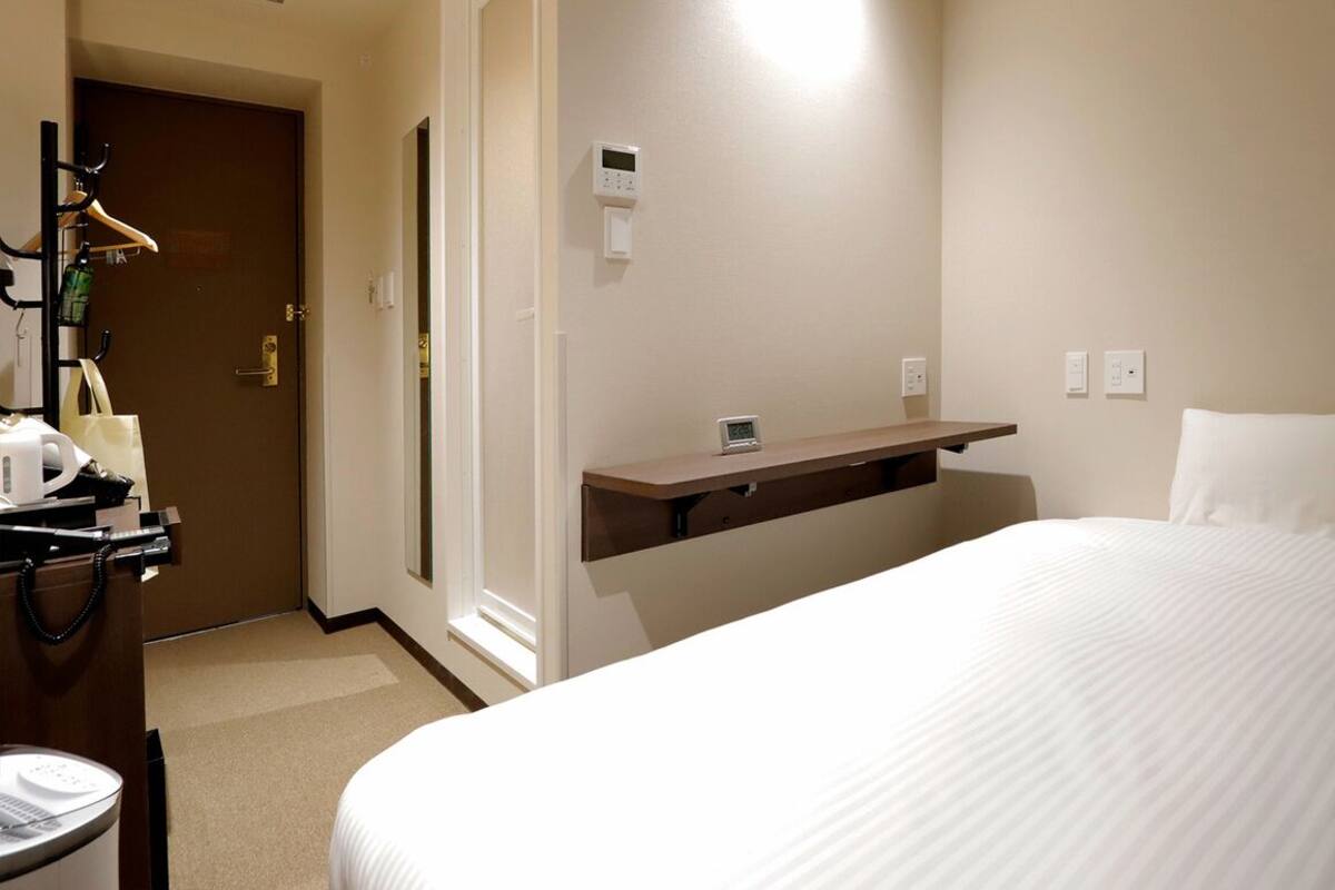 A hotel room with a bed, a desk with a chair, a coat rack, and a door.