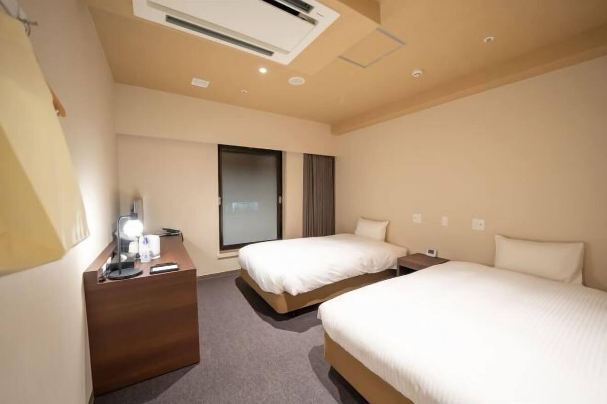 A hotel room with a desk, two beds, a window, and an air conditioning unit.