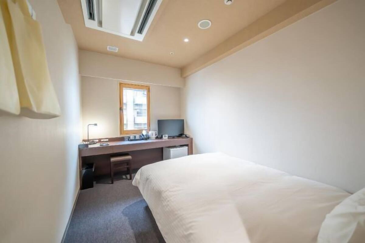 A hotel room with a bed, a desk with a computer, a chair, a window, and a ceiling vent.