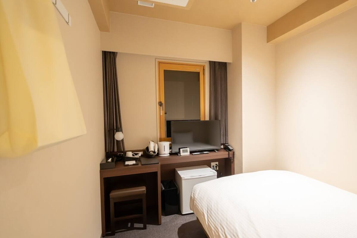 A hotel room with a bed, a desk with a computer, a telephone, and a small stool.