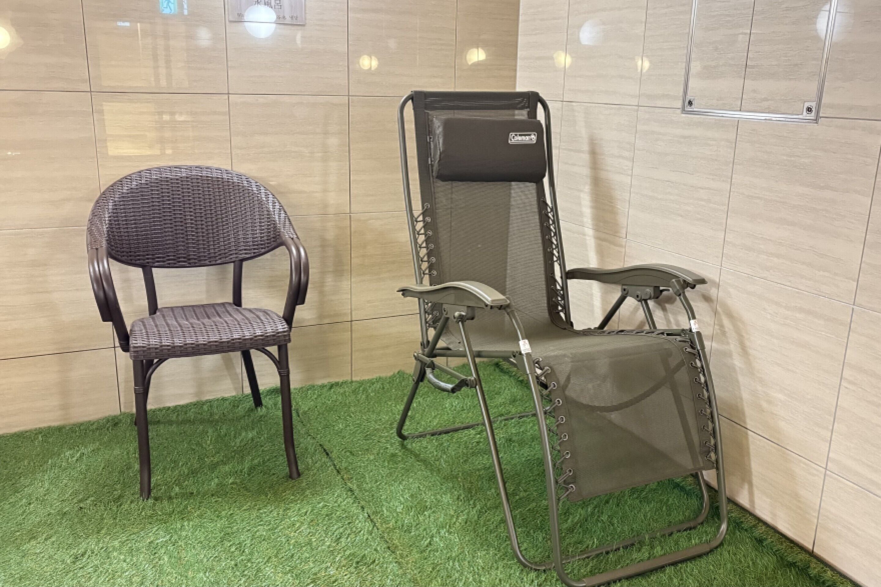 Ein Zimmer mit einem Korbstuhl, einem Klappstuhl mit Netzrücklehne und einem Boden aus grünem Kunstgrün.