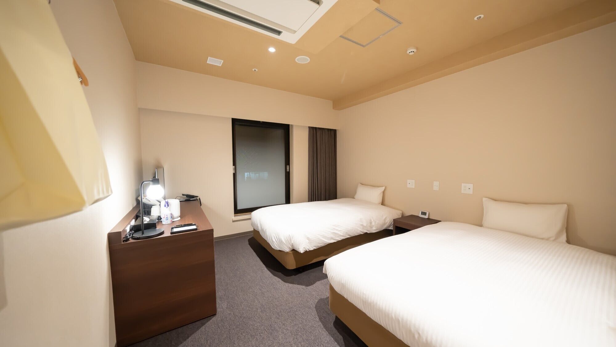 A hotel room with two beds, a desk with a lamp, a window with curtains, and a ceiling light.