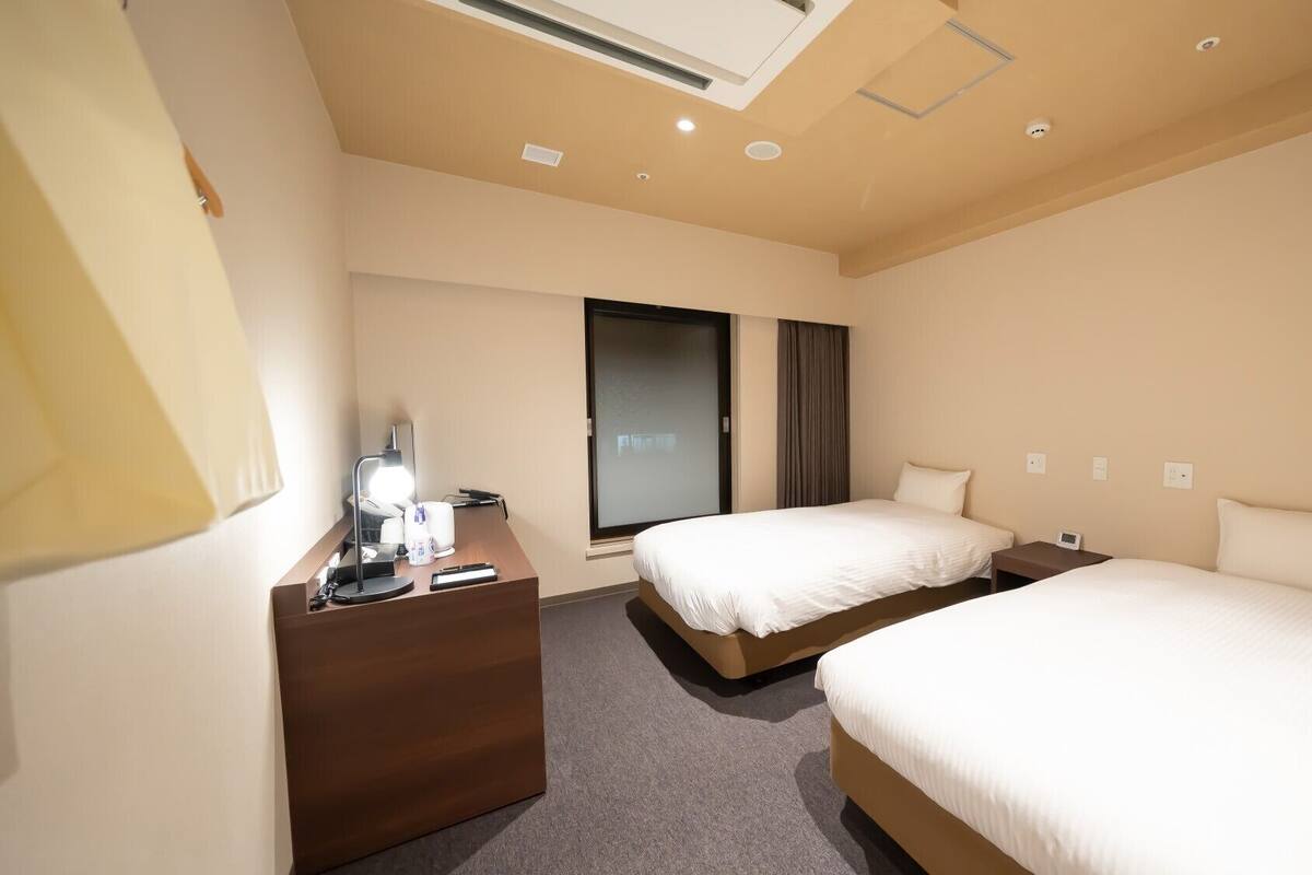 A hotel room with two beds, a desk with a lamp, a window with curtains, and a ceiling light.