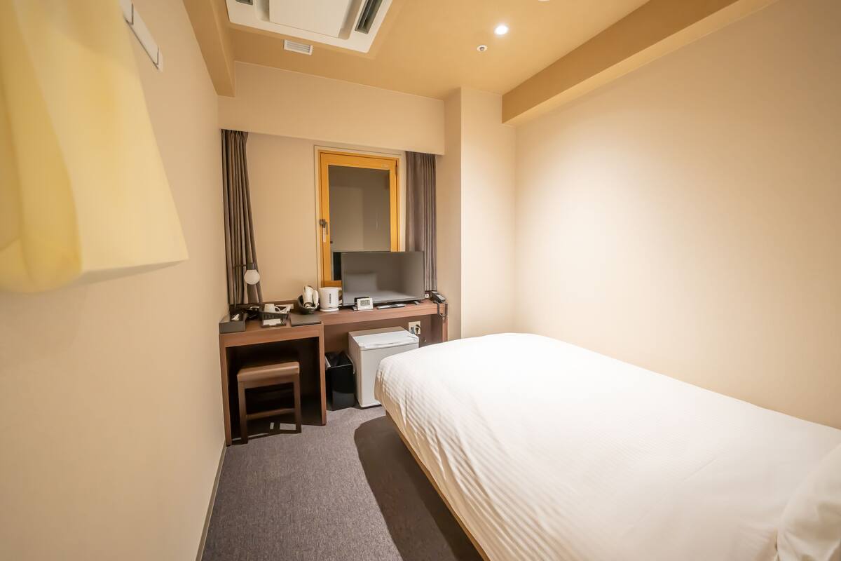 A hotel room with a bed, a desk with a computer, a chair, a small table, a mirror, and a window with curtains.