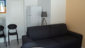 Un espace de vie comprenant un canapé foncé, une petite table ronde, deux chaises, une porte blanche, un réfrigérateur blanc et un lampadaire.