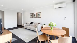 Een moderne woonkamer met een bank, een salontafel en een eethoek met stoelen. Er is een flatscreen televisie, een bijzettafeltje en een vaas met bloemen.