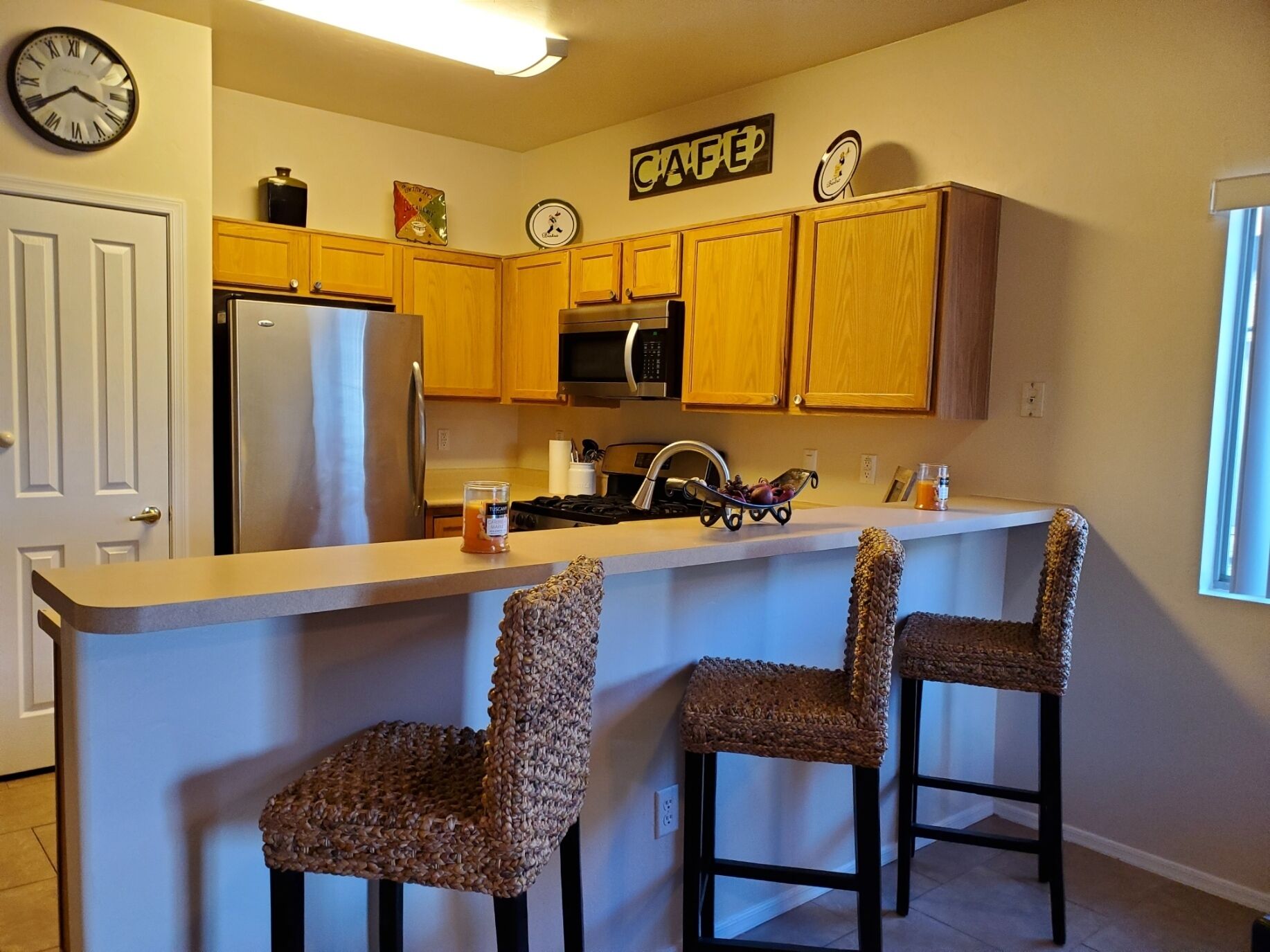 A kitchen with wooden cabinets, a stainless steel refrigerator, a microwave, and a coffee station. There are two woven chairs at the counter.