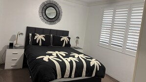 A bedroom with a bed, a nightstand, a lamp, a ceiling fan, a window with blinds, and a wall-mounted mirror. - MANLYS LITTLE SECRET (Manly)