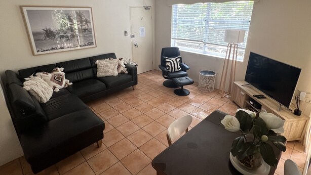 Ein Wohnzimmer mit einem schwarzen Ecksofa, einem Esstisch mit Stühlen, einem Deckenventilator, einem Fernseher und einem Fenster mit Jalousien.