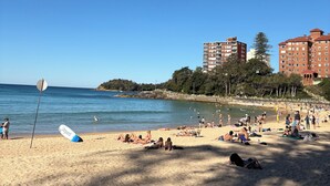 Ein Strand mit badenden Personen, einem Rettungsschwimmerstand und Gebäuden im Hintergrund.