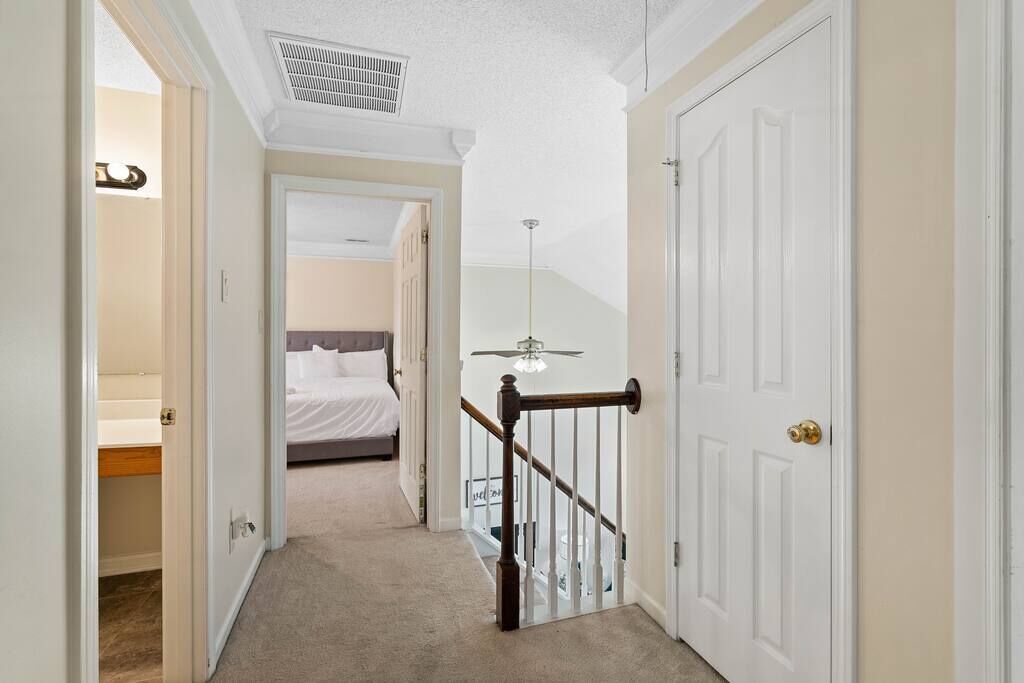 A hallway with a carpeted floor, a wooden railing, and a bed in the background.
