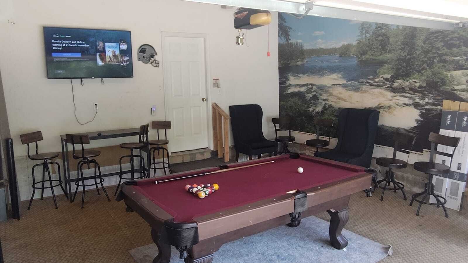 A pool table with chairs, a television displaying sports news, and a mural of a river scene.