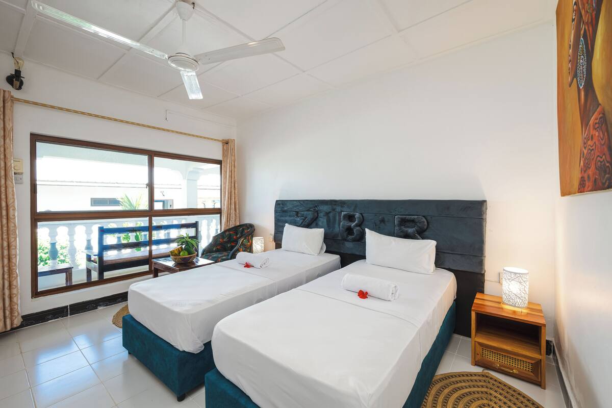 A hotel room with two beds, a ceiling fan, a large window with a view, and a small table with a lamp.