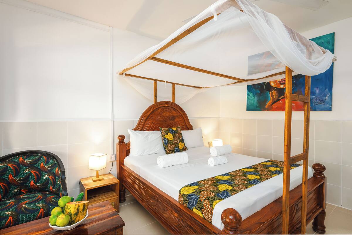 A four-poster bed with a canopy, a wooden nightstand with a lamp, a chair with a colorful cushion, and a bowl of fruit on the nightstand.