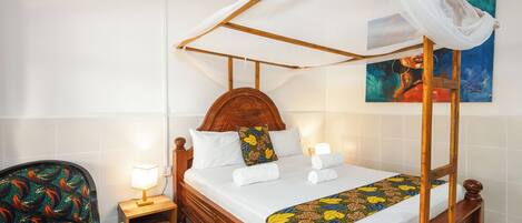 Un lit à baldaquin, une table de chevet en bois avec une lampe, une chaise avec un coussin coloré et un bol de fruits sur la table de chevet.