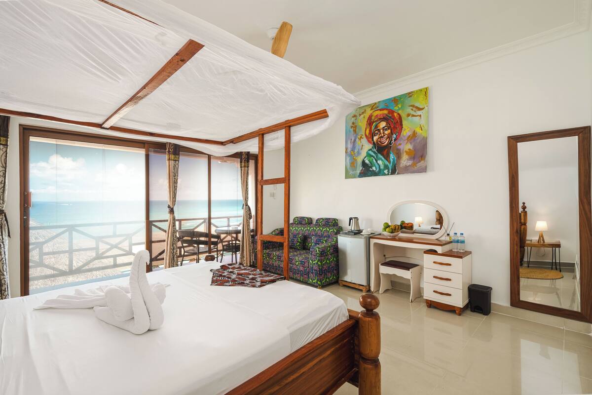 A bedroom with a bed, a desk, a chair, a dresser, a painting, and a view of the ocean.