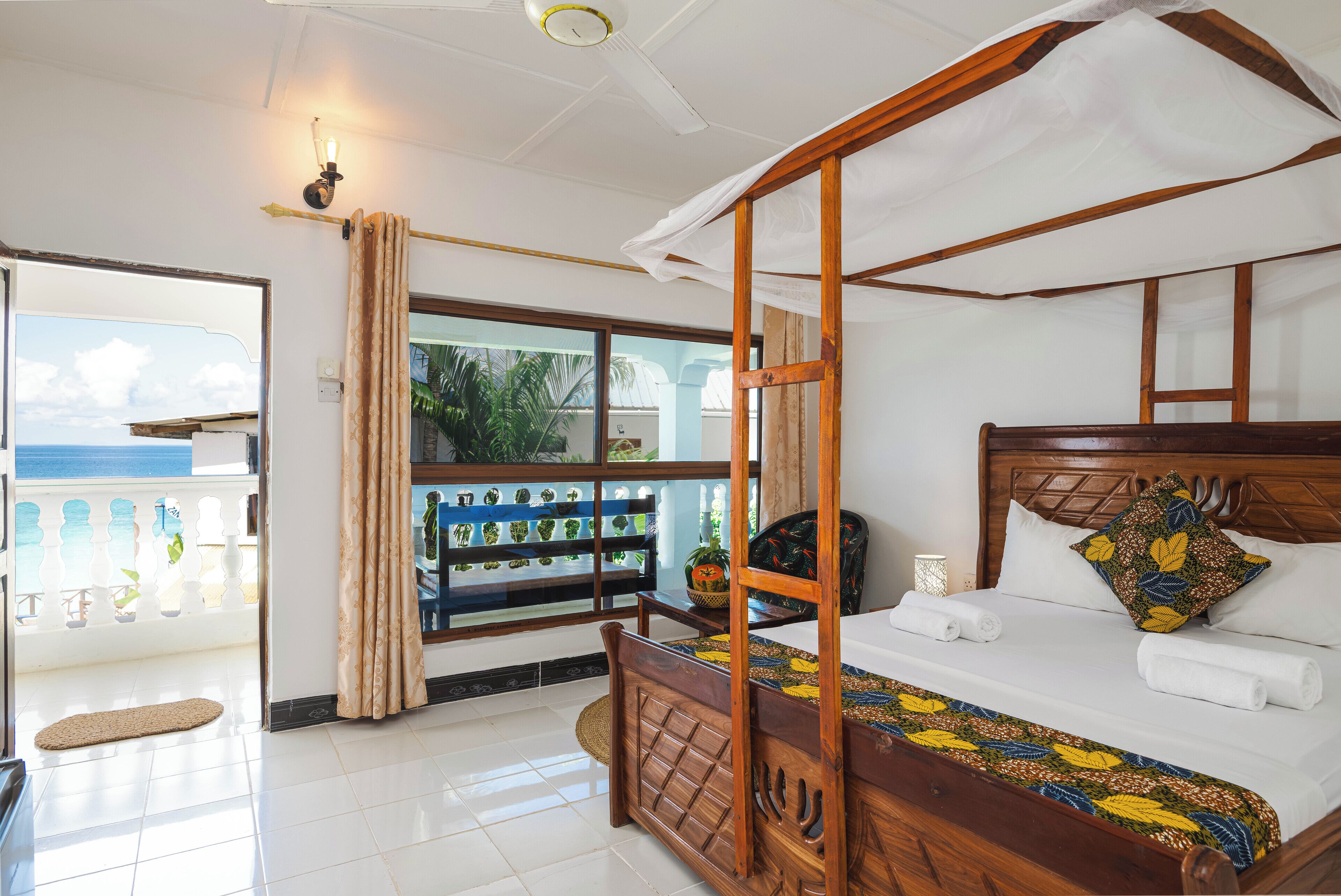 Een hemelbed met een bedovertrek met dessin, een houten hoofdbord en uitzicht op de zee vanaf een balkon met potplanten.