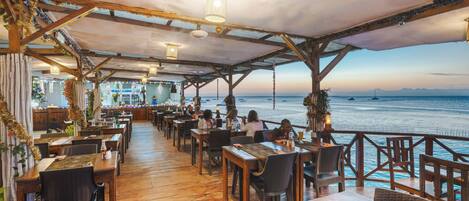 Un restaurante al aire libre con mesas y sillas de madera, vista al mar y un área cubierta con luces colgantes.