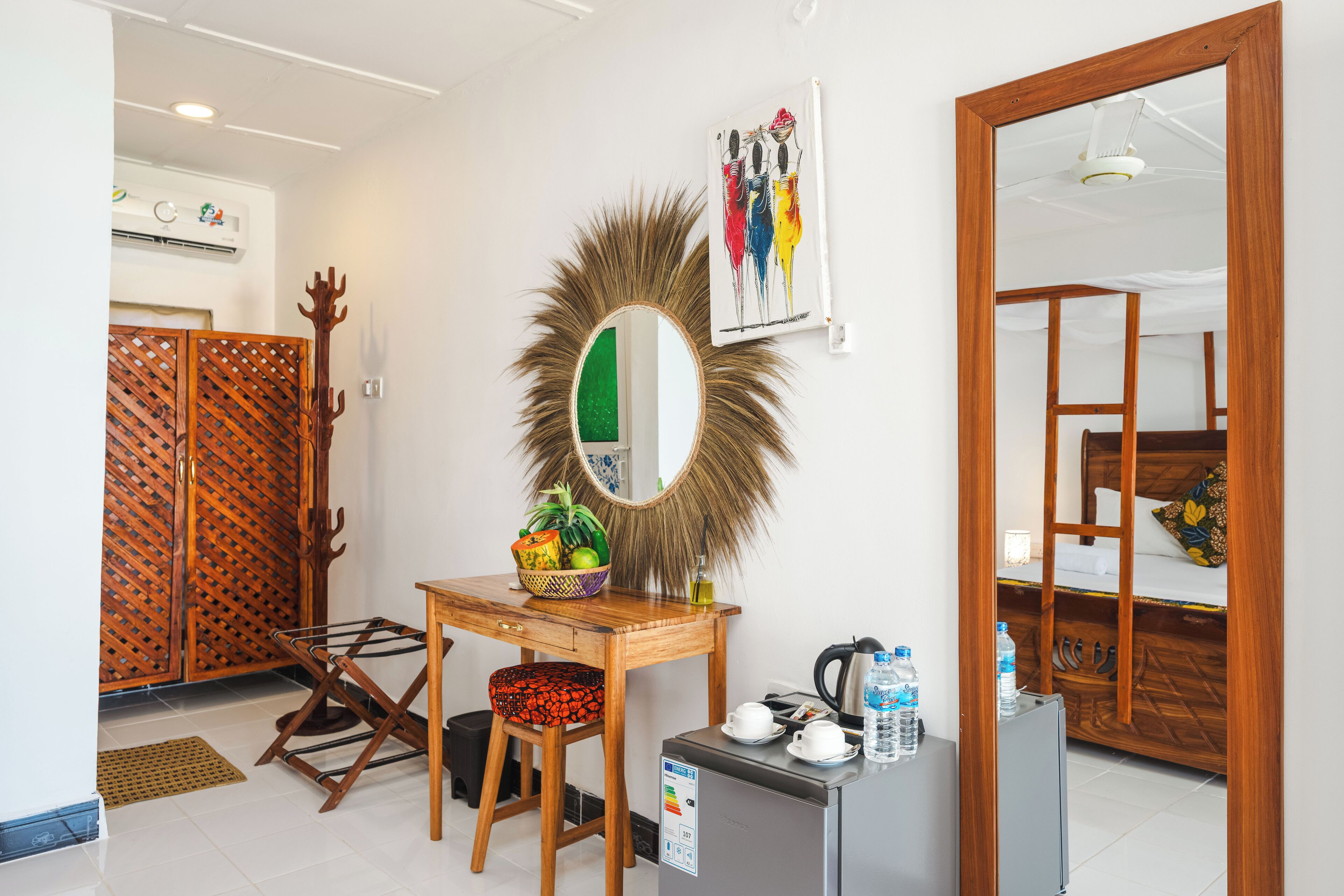 A hotel room with a wooden desk, a mirror, a small table with a basket of fruit, a chair, a mini fridge, and a bed with a wooden headboard.
