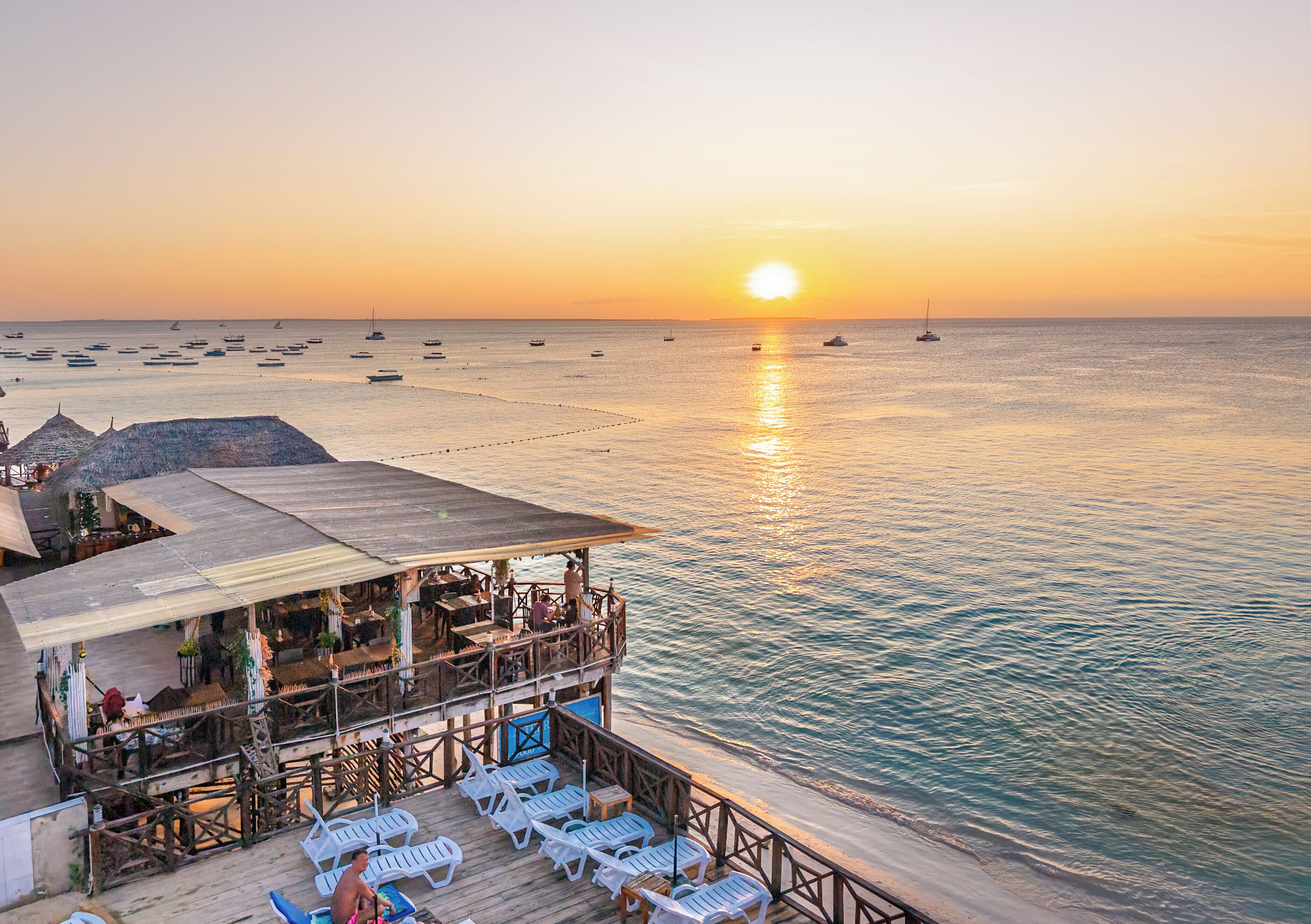 A seaside restaurant with outdoor seating overlooking the ocean, boats in the distance, and a sunset.