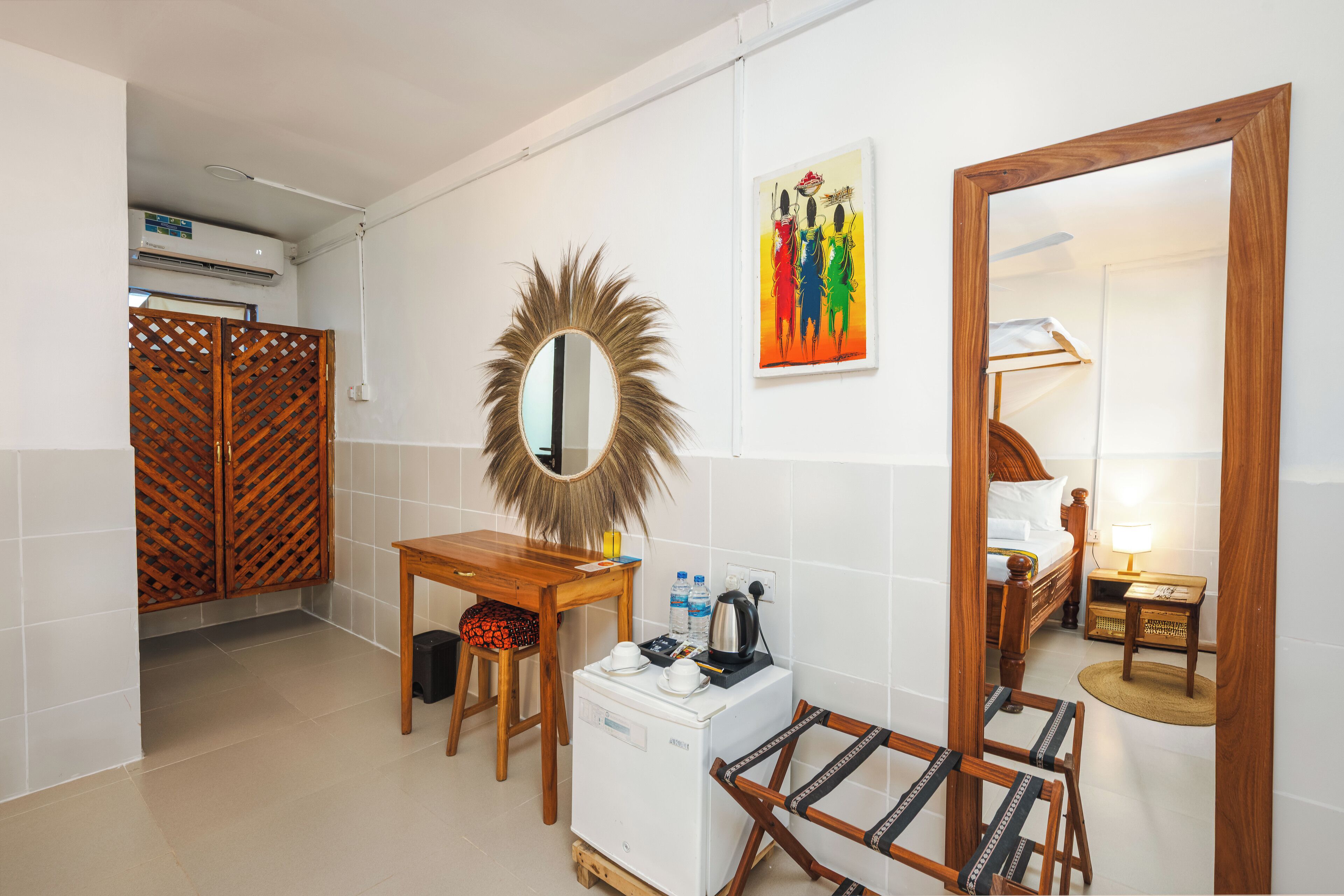 Ein kleines Hotelzimmer mit einem hölzernen Bett, einem kleinen Schreibtisch, einem Kühlschrank, einem Spiegel und einem farbenfrohen Kunstwerk an der Wand.
