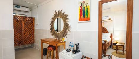 A compact hotel room with a wooden bed, a small desk, a refrigerator, a mirror, and a colorful artwork on the wall.