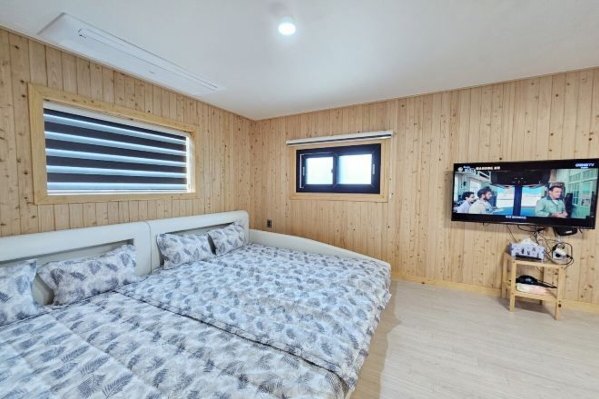A bedroom with a large bed, wooden walls, two windows, and a TV on a wooden stand.