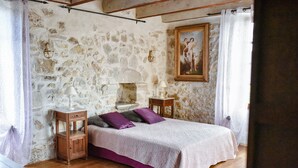 A bedroom with a stone wall, a bed with a white and purple bedspread, two bedside tables with lamps, and a painting on the wall. - Magnificent private Mas Ardechois with swimming pool (Labastide-de-Virac)