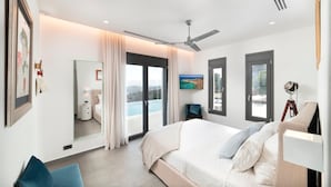 Een moderne slaapkamer met een groot bed, een plafondventilator, een televisie en uitzicht op zee door de ramen.