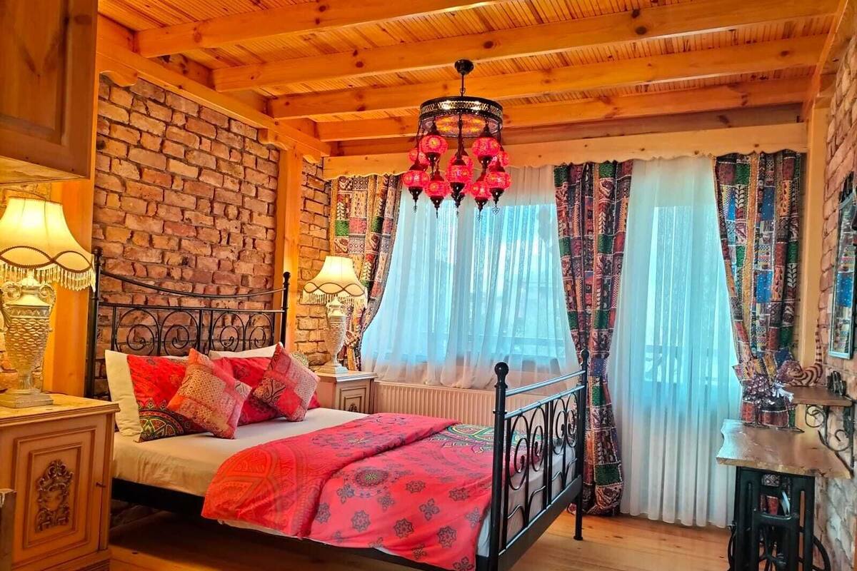 Ein gemütliches Schlafzimmer mit Backsteinwand, Holzboden und einem Bett mit gemusterten Betttextilien. Ein Nachttisch mit Lampe und ein Fenster mit transparenten Vorhängen.
