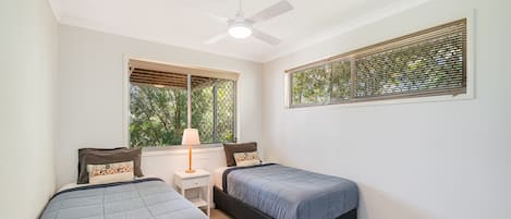 A bedroom with two beds, a ceiling fan, a nightstand with a lamp, and a window with blinds.