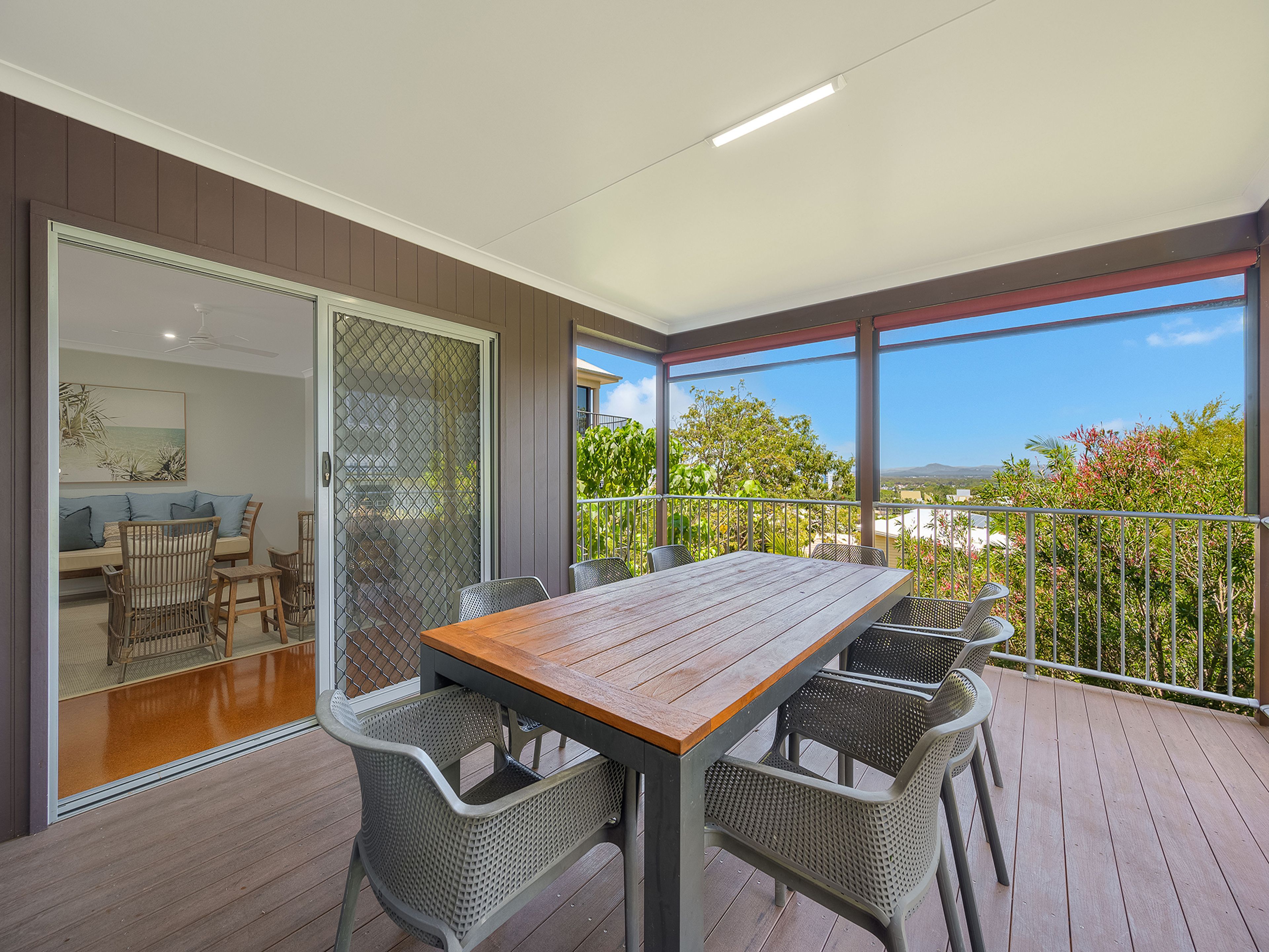 A wooden deck with a table and chairs, a sliding glass door leading to a balcony, and a view of greenery and buildings.