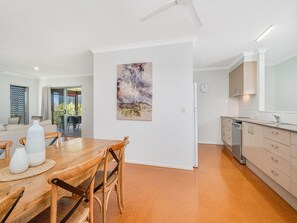 A modern kitchen with a dishwasher, a dining area with a wooden table and chairs, and a large painting on the wall. - SaltyS Place, Pet Friendly Escape (CALOUNDRA)