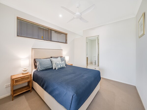Quarto com cama, mesa de cabeceira, abajur, ventoinha no teto e porta de banheiro.