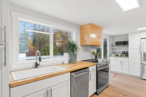 Une cuisine moderne avec des armoires blanches, un comptoir en bois, des appareils électroménagers en acier inoxydable et une grande fenêtre donnant sur la forêt.