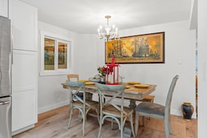 A dining area with a wooden table, four chairs, a chandelier, and a large painting on the wall.