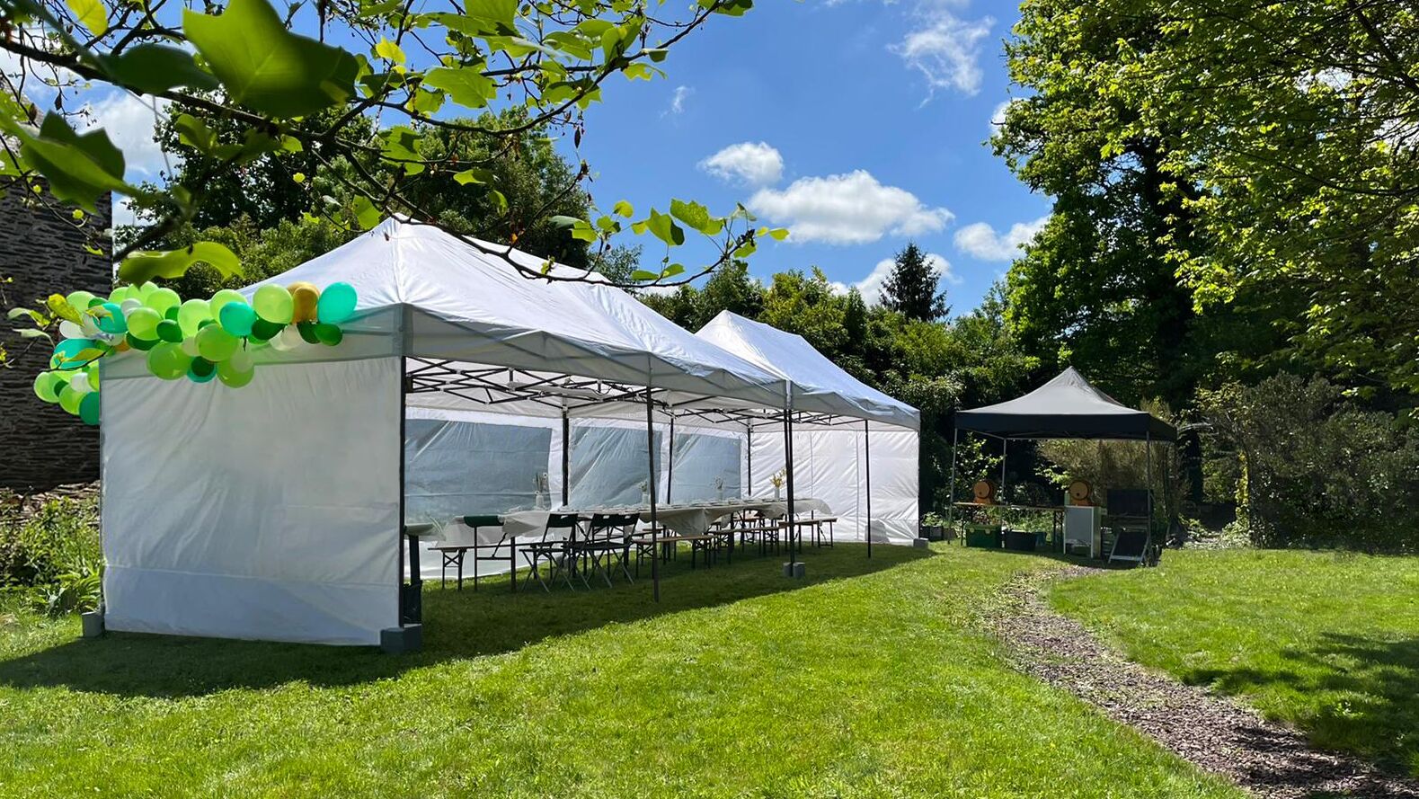 A white tent with tables and chairs set up outdoors, surrounded by greenery and trees.