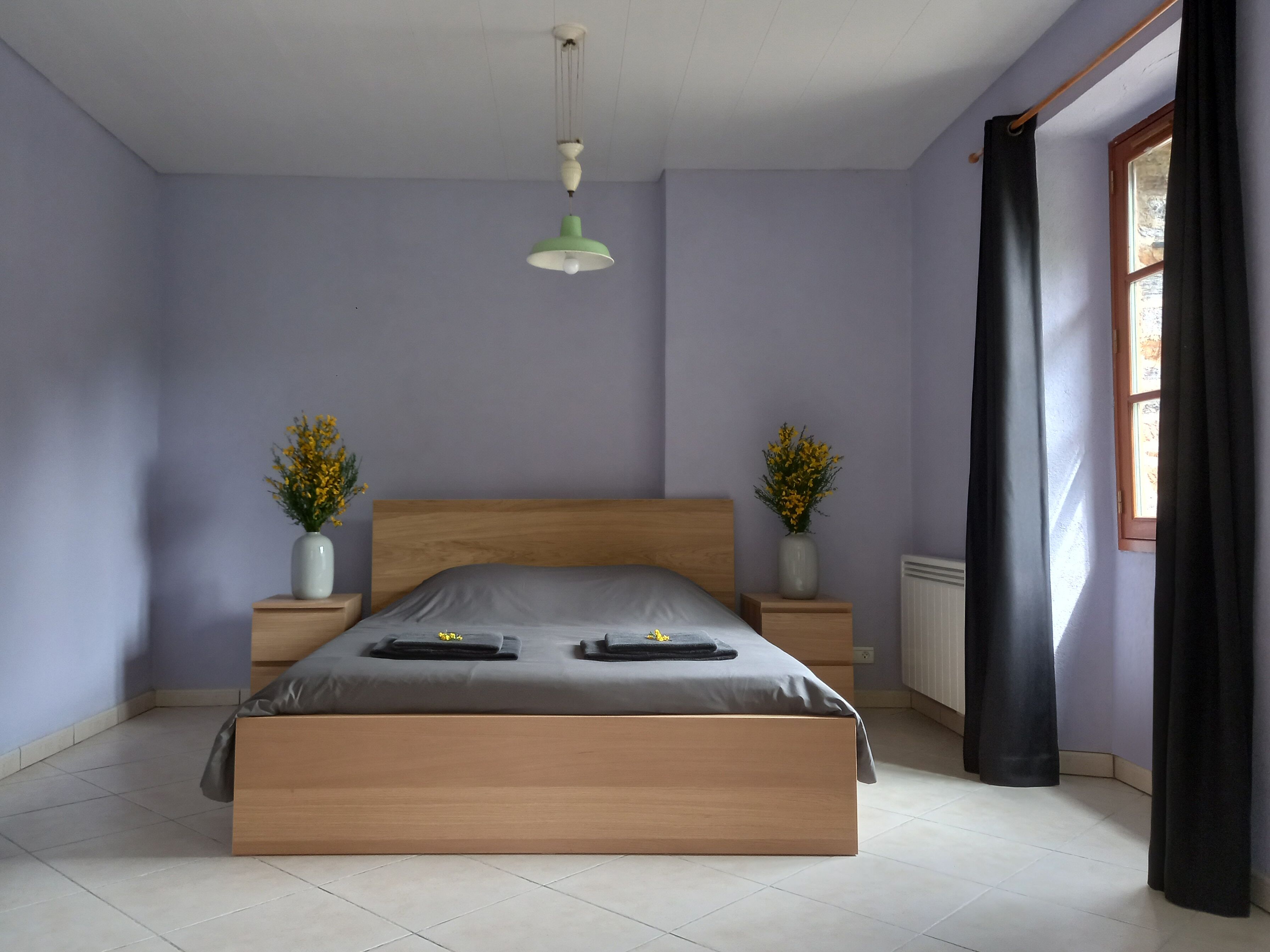 A bedroom with a wooden bed, two bedside tables with vases, a hanging light fixture, and a window with curtains.