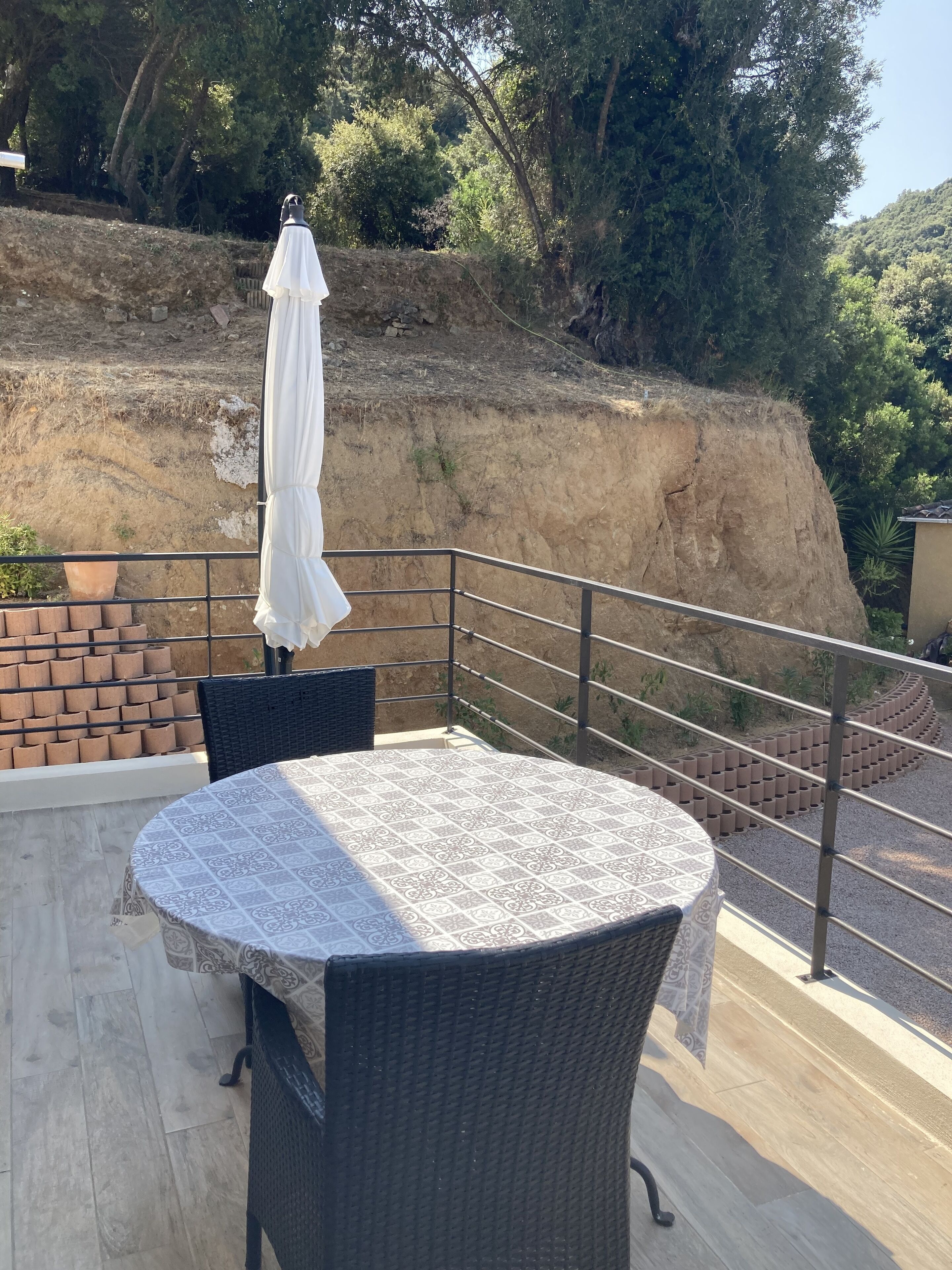 A balcony with a round table and chairs, a white umbrella, and a view of a hillside.