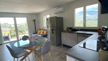 A modern kitchen with a built-in refrigerator, a dining area with a glass table and chairs, and a view of a mountainous landscape through large windows.