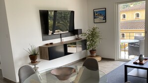 Una sala de estar moderna con una mesa de comedor de vidrio, sillas tapizadas en gris, un televisor de pantalla plana montado en la pared y vistas de un edificio a través de una puerta de cristal.