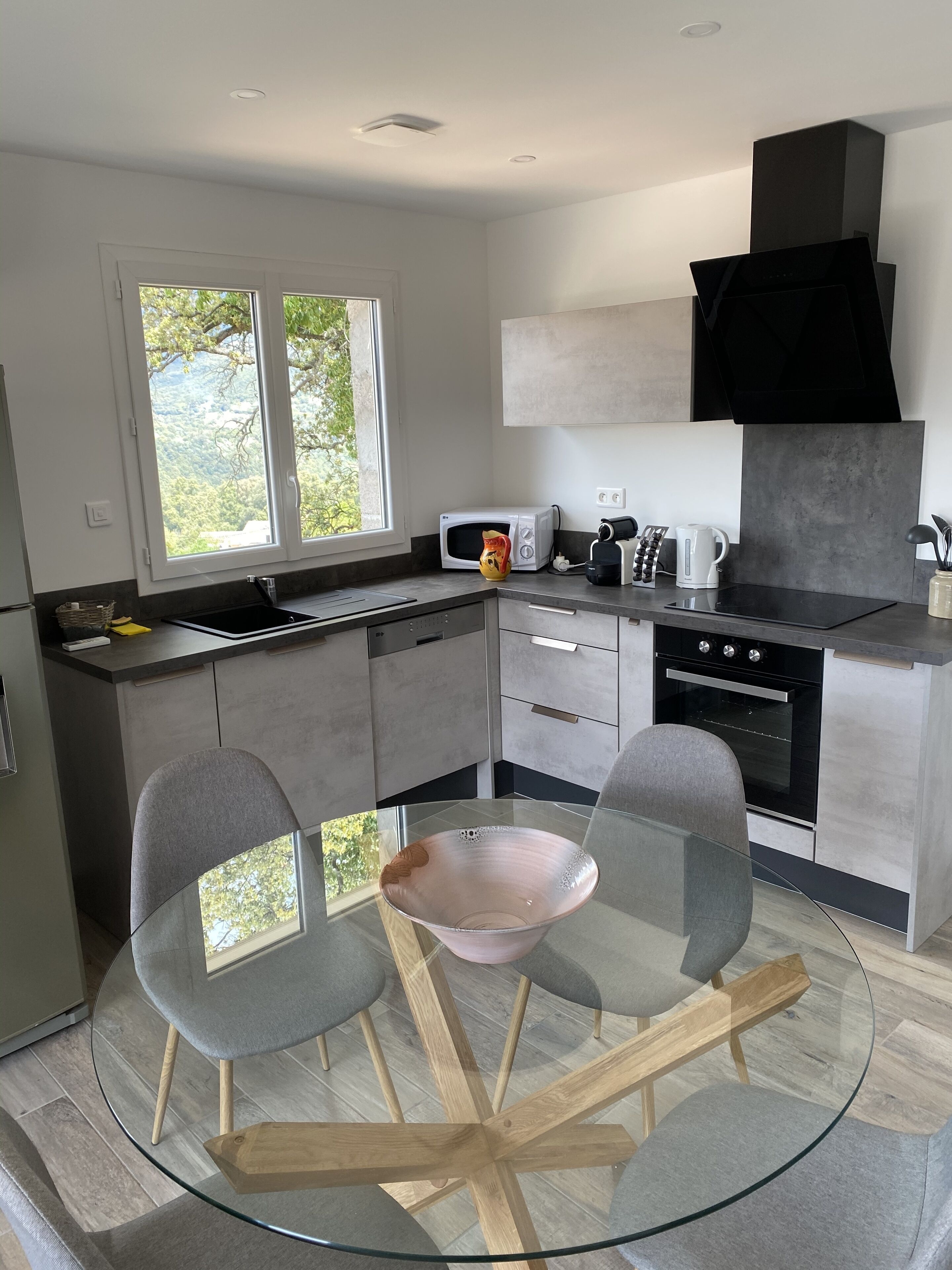 A modern kitchen with a glass dining table, grey chairs, a microwave, kettle, and a large window offering a view of greenery.