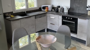 Una cocina moderna con una mesa de comedor de vidrio, sillas grises, un microondas, hervidor y un amplio ventanal con vistas a la vegetación.
