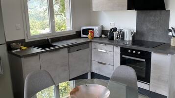 A modern kitchen with a glass dining table, grey chairs, a microwave, kettle, and a large window offering a view of greenery.