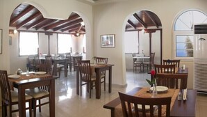 A spacious dining area with multiple wooden tables and chairs, large windows, and a high ceiling with wooden beams. - Kassandra Bay (Kassandra)