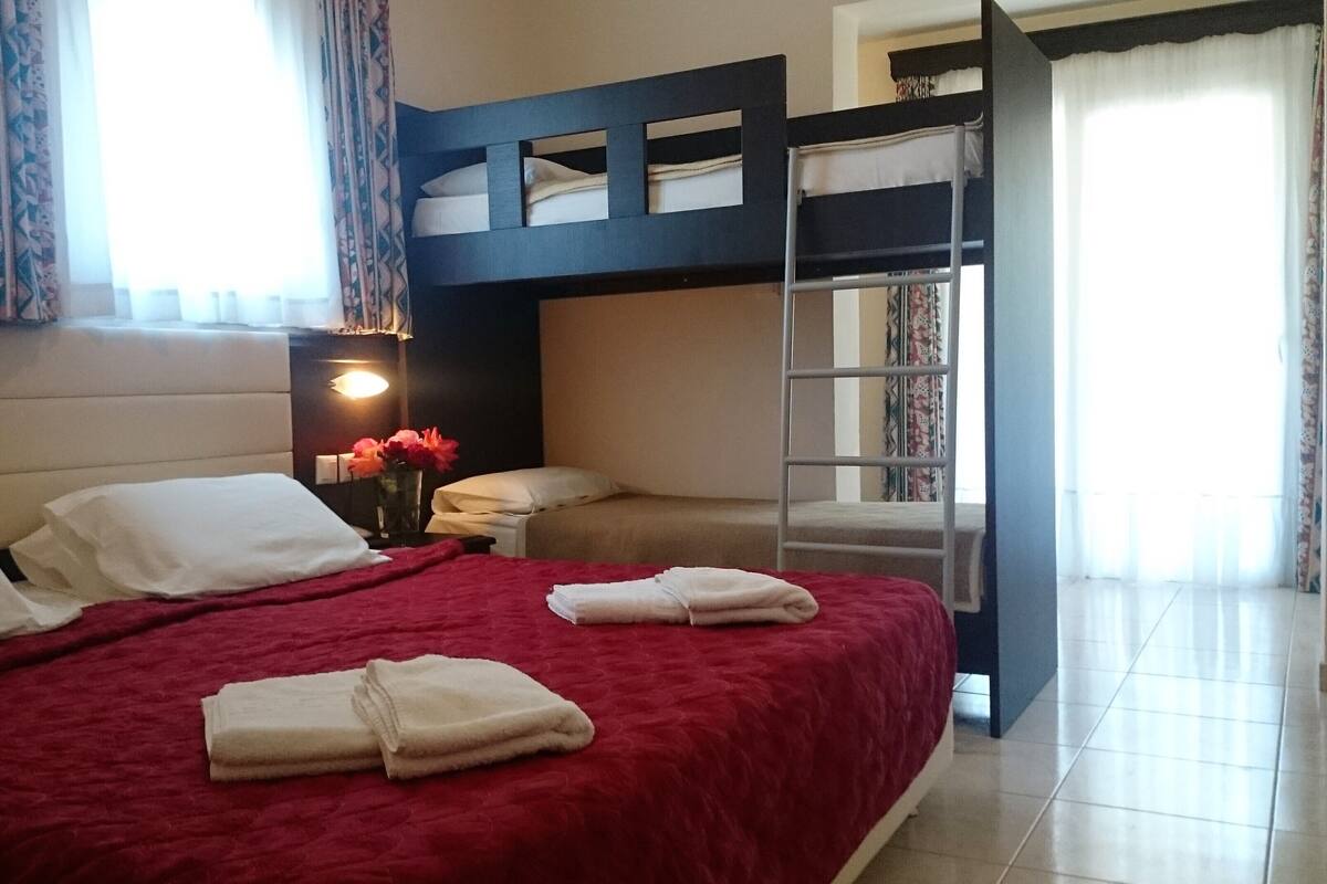 A hotel room with a bunk bed, a red bedspread, white pillows, a bedside table with a lamp, and a vase with flowers.