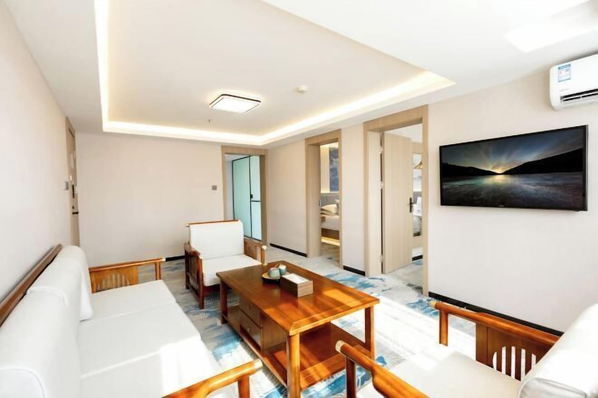 A modern hotel room with a wooden dining table, white sofa, and a large window.