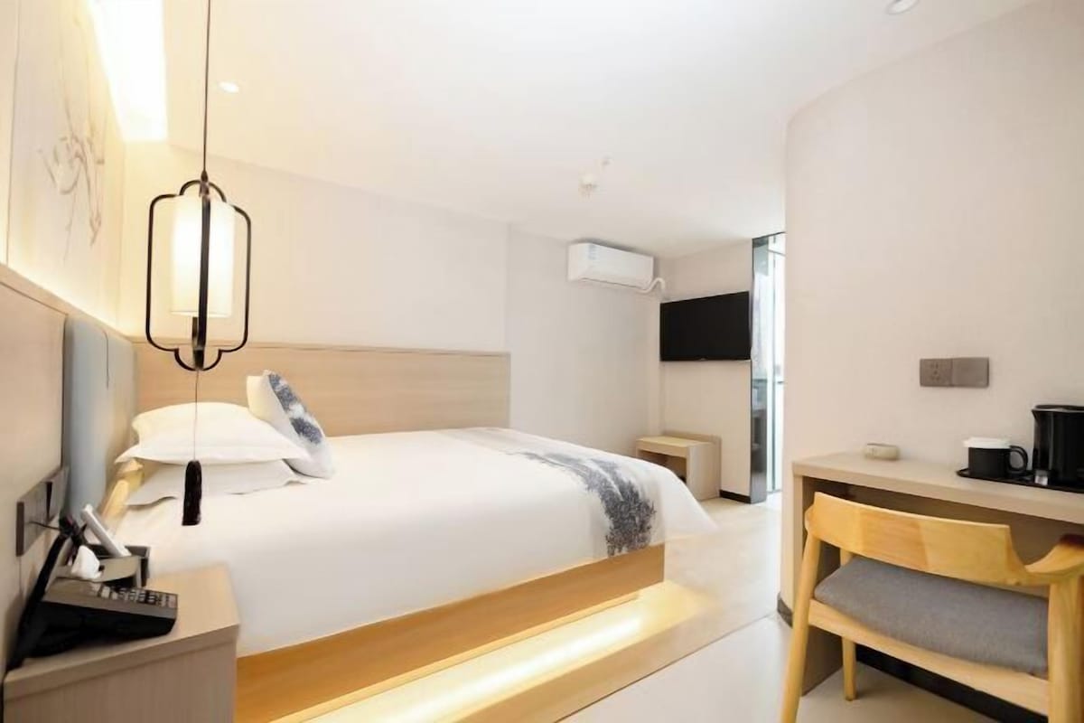 A modern hotel room with a large bed, bedside table, and a desk with a chair.