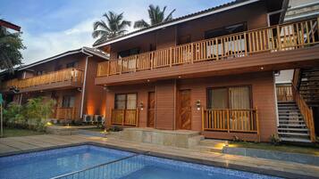 Un hotel de dos pisos con piscina, balcones de madera y un jardín tropical.